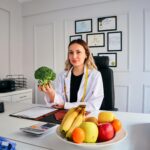 Female nutritionist in office holding broccoli, surrounded by fruits, promoting healthy lifestyle.